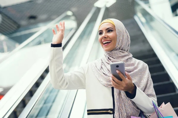 Arab woman walking in shopping mall with shopping bags and using smartphone