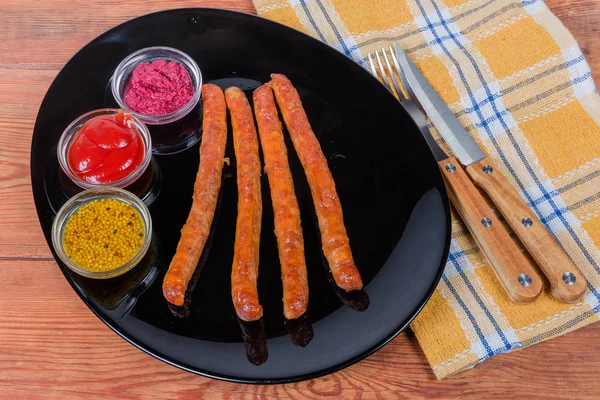 Embutidos de cerdo al horno en plato negro, varios condimentos, tenedor, cuchillo — Foto de Stock