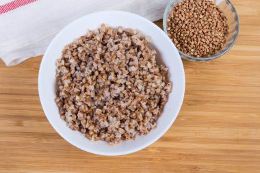 Prepared buckwheat porridge in white bowl and uncooked buckwheat groat in small glass bowl on a wooden surface, top view clipart