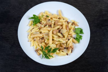 Kaynamış silindir şeklinde makarna, kızarmış mantar ve soğanla karıştırılmış siyah yüzeyde beyaz tabakta.