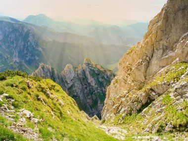 Dik kayalık çıkıntıları olan derin dağ geçidi güneş ışığında uzak tepelere karşı ön planda.