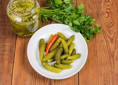 Turşulu minik salatalıklar da beyaz tabakta turşu olarak adlandırılır ve küçük cam bir kavanozda, kırsal masada taze maydanoz, üst manzara.