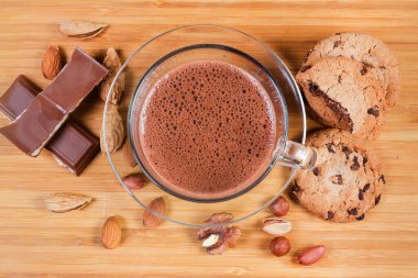 Çikolata parçalı kurabiyelerin, çikolata parçalarının ve ahşap yüzeyde çeşitli fındıkların arasında tabağı olan cam bardakta köpüklü sıcak çikolata.