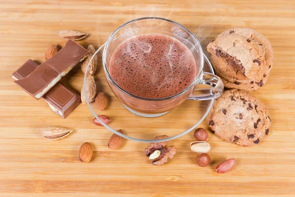 チョコレートチップクッキー チョコレートピース 木製の表面上の様々なナッツの間でソーサーとガラスカップで泡ホットチョコレート — ストック写真