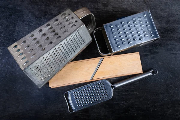 Old kitchen box graters with multiple grating surfaces and different holes, flat grater and wooden grater with special stainless steel blade on the black surface