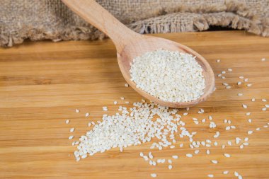 Tahta kaşıkta beyaz susam tohumları ve ahşap yüzeyin yanına serpilmiş, seçici bir odak noktasında yakın plan