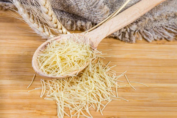 Uncooked very thin short-cut round section pasta known as Filini pasta in the wooden spoon and scatterd beside, wheat ears on wooden surface with burlap