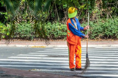 Turuncu ceketli, mavi ceketli, turuncu pantolonlu ve elinde süpürgeyle sarı şapkalı orta yaşlı bir adam yolu süpürüyor.