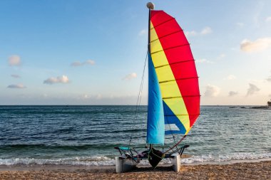 Kırmızı, sarı ve mavi yelkenli ve ekipmanlı güzel beyaz katamaran, bulutlarla kaplı, turkuaz deniz kıyısında, ıslak kumsalda, altın plajda.