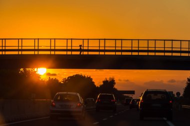Güzel bir gün batımı, altın güneş diski, kırmızı bulutlar, köprünün siyah bir silueti ve yolda bir kız.