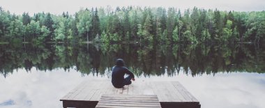 Adam güzel bir gölün iskelesinde oturuyor, panoramik bir fotoğraf.