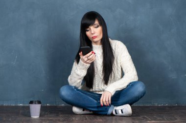 Modaya uygun esmer kız yerde oturuyor, akıllı telefon ekranına bakıyor, üzgün. Monokrom mavisi, modaya uygun eski püskü bir arkaplan üzerindeki stüdyo portresi, metin için alan.