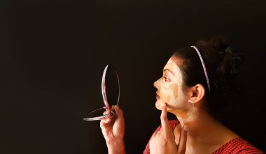 an indian lady with face pack applied in half dried condition checking her face in a mirror clipart