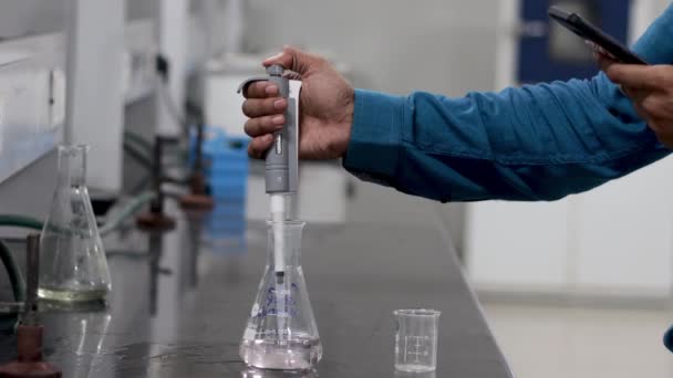 Handen van een onderzoeker die een kleurloze chemische stof met behulp van een microfoon uit een erlenmeyer in een bekerglas giet — Stockvideo