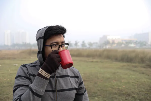 Un uomo che beve caffè caldo da una tazza rossa e vestito con indumenti invernali e guanti con particolare attenzione ai modelli faccia con una nebbia foschia sullo sfondo — Foto Stock