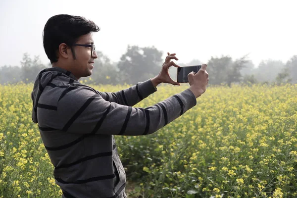 A man in hoodie and spectacles clicking nature photo with his smartphone in a mustard field with yellow blossomed mustard flowers during day time — 스톡 사진