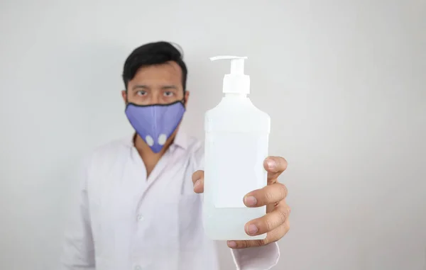 Un medico maschio in maschera e mantello bianco con una bottiglia bianca di disinfettante per le mani isolata su sfondo grigio con spazio per il testo — Foto Stock