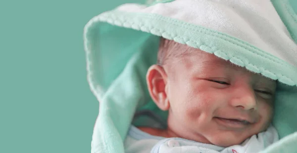Ένα χαμογελαστό νεογέννητο μωρό με λακκάκι στο μάγουλο τυλιγμένο σε πράσινη πετσέτα θαλάσσης με κουκούλα με μάτια κλειστά με επιλεκτική εστίαση στο μπροστινό μάτι — Φωτογραφία Αρχείου