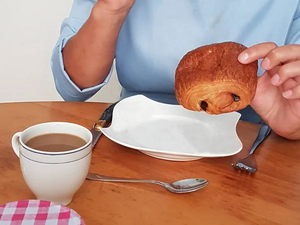 A girl in a blue dress at a table eating a traditional French breakfast: a croissant and coffee.