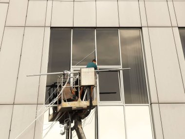 Bir kaldırma platformundaki iki işçi yüksek bir binanın dışındaki bir pencereyi tamir ediyorlar. Gökdelenlerin camlarını siliyorum. Yüksek irtifa çalışması tehlikesi. Gökdelenlerin onarımı ve bakımı