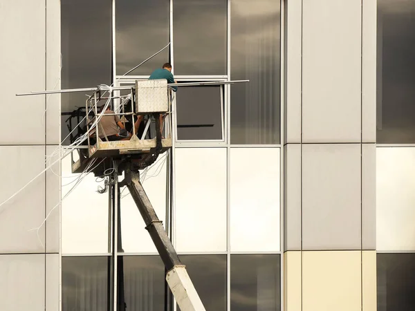 Dva dělníci na zvedací plošině opravují okno na vnější straně vysoké budovy. Mytí okna mrakodrapů. Nebezpečí práce ve velké výšce. Údržba a opravy mrakodrapů — Stock fotografie