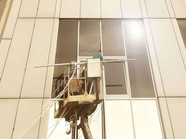 Bir kaldırma platformundaki iki işçi yüksek bir binanın dışındaki bir pencereyi tamir ediyorlar. Gökdelenlerin camlarını siliyorum. Yüksek irtifa çalışması tehlikesi. Gökdelenlerin onarımı ve bakımı — Stok fotoğraf
