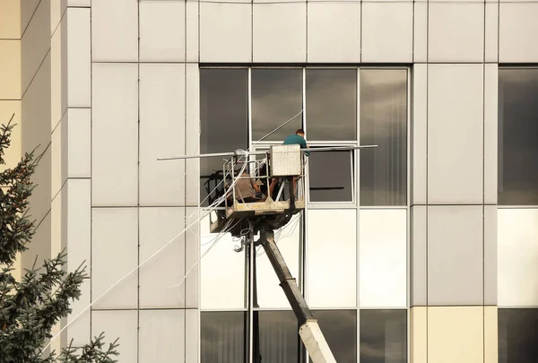 Dois trabalhadores numa plataforma de elevação estão a reparar uma janela do lado de fora de um edifício alto. A lavar uma janela de arranha-céus. Perigo de trabalho de alta altitude. Manutenção e reparação de arranha-céus — Fotografia de Stock