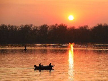 İki balıkçı, bir erkek ve bir kadın, turuncu bir günbatımının arka planında nehirde bir motorlu teknede yüzerler. Aşıklar için romantik bir ortam. Akşam balıkçılığı. Suyla seyahat et. Doğal bir fenomen