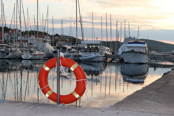 橙色生活圆环在码头在克罗地亚码头反对风帆游艇的背景。安全的水和拯救溺水。海港的设备。救援队的装备 — 图库照片