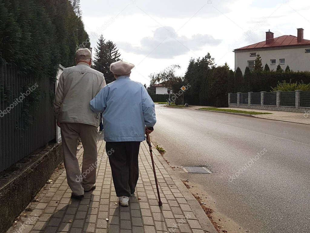 A pair of elderly people walk along the sidewalk along the road holding hands. Grandfather and grandmother on a walk in a residential area. Movement health of the elderly. Happy old age