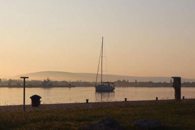 Yelkenli yat, sabah güneşinin altında marinadaki motorun altına girer. Şafak limanda. Deniz fenerine karşı iskelede boş bir bank. Dönüşün ve beklemenin sembolleri. Sakin bir romantik. 