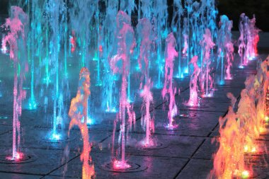 Multi-colored water jets of the musical fountain in the evening city. Place of rest and relaxation. The colors of the rainbow in the spray. Illumination of night streets and recreation. Decoration