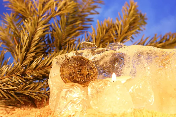 Neujahrsdekoration. Bitcoin versank im Eis und wurde von der Flamme einer roten Kerze vor dem Hintergrund von Christbaumzweigen und blauem Himmel aufgehellt. das Einfrieren von Finanzvermögen — Stockfoto