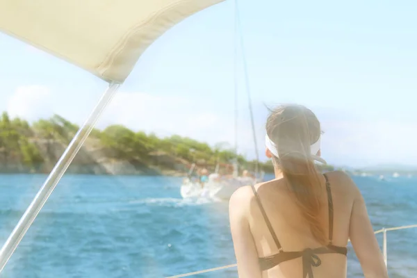 A young slender girl in a swimsuit sits on the stern and watches a sailing yacht passing by. A luxury holiday on the water for wealthy people. Prestigious lifestyle. Vacation and summer sea voyage