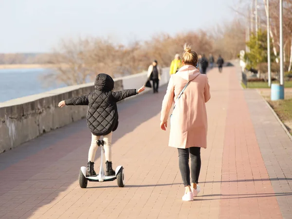 Seorang ibu muda dan anak perempuan berjalan di sepanjang promenade. Seorang gadis mengendarai taman dengan skuter gyro. Modern mode ekologi listrik transportasi. Ekologi di kota. Aktivitas anak-anak. Olahraga untuk kesehatan — Stok Foto