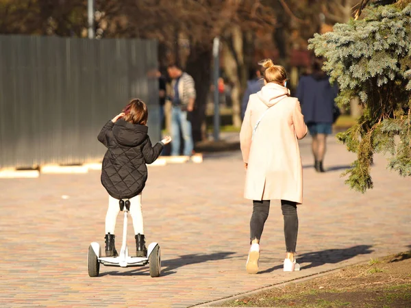 Seorang ibu muda dan anak perempuan berjalan di sepanjang promenade. Seorang gadis mengendarai taman dengan skuter gyro. Modern mode ekologi listrik transportasi. Ekologi di kota. Aktivitas anak-anak. Olahraga untuk kesehatan — Stok Foto