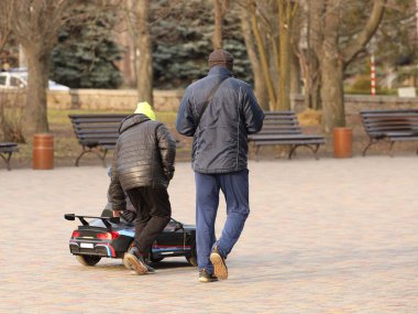 Bir adam ve oğlu güneşli bir bahar gününde parkta elektrikli bir arabada küçük bir çocuğa biniyorlar. Çocukların ahenkli gelişimi için etkinlik. Modern yüksek teknoloji oyuncaklar. Baba oğul yetiştiriyor.