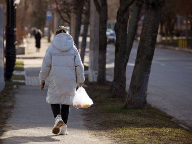 Elinde bir paket olan bir kadın gün ışığında kaldırımda yürüyor. Şehir caddesinde yaya. Evdeki bir kadın metresin günlük hayatı. Markete alışveriş gezisi. Plastik torba kullanıyor.
