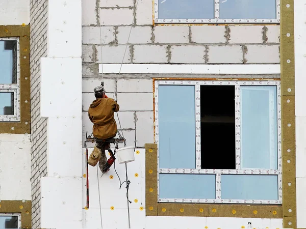 A builder in climbing equipment insulates the facade of a high-rise building with foam plastic. Safety measures for high-altitude construction work. Heat conservation in homes and energy savings