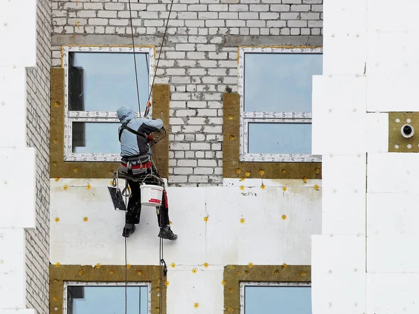 A builder in climbing equipment insulates the facade of a high-rise building with foam plastic. Safety measures for high-altitude construction work. Heat conservation in homes and energy savings