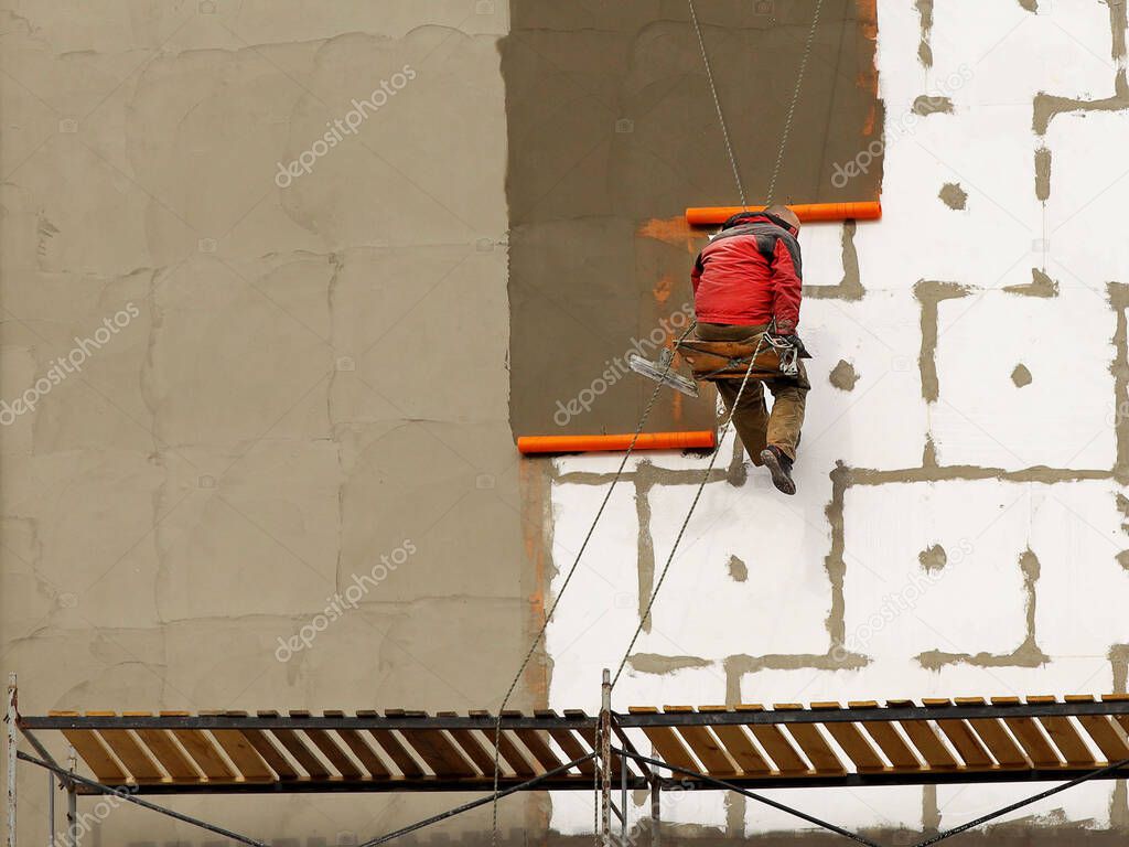A builder in climbing equipment insulates the facade of a high-rise building with foam plastic. Safety measures for high-altitude construction work. Heat conservation in homes and energy savings.