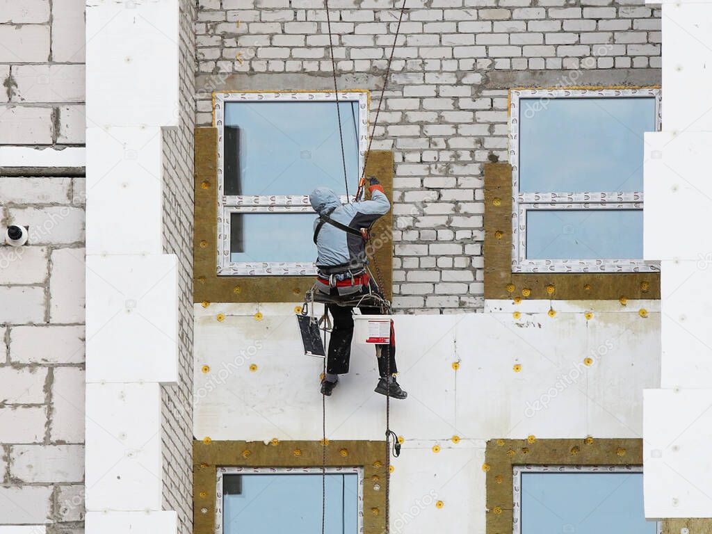 A builder in climbing equipment insulates the facade of a high-rise building with foam plastic. Safety measures for high-altitude construction work. Heat conservation in homes and energy savings.