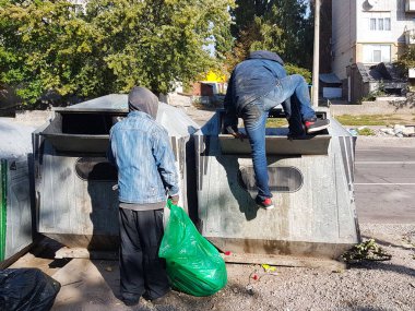 Homeless people seek food and valuables in a trash can. The disadvantaged louis carry a beggarly lifestyle. Sorting municipal waste. Violation of human rights and dignity. Tramps in the city. clipart