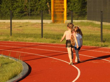 Futbol sahasının etrafındaki stadyumun koşu bandında koşan iki kız. Spor kompleksinin yapay kapağı. Sağlıklı faaliyet içinde ebeveynlik. Nüfusta mevsimsel soğuk algınlığının önlenmesi. Açık hava..