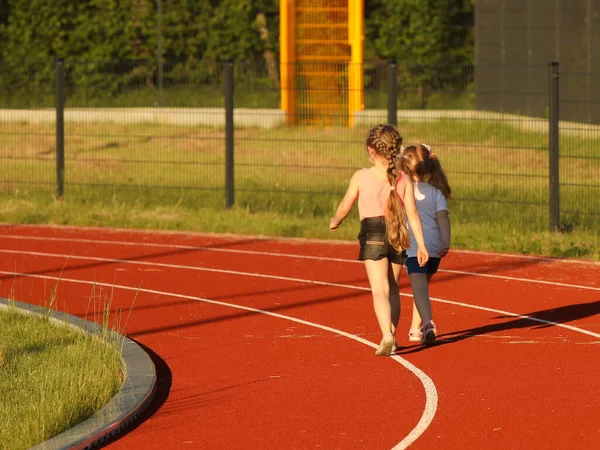 Two girls run on the treadmill of the stadium around the football field. Artificial cover of the sports complex. Parenting in healthy activity. Prevention of seasonal colds in the population. Outdoor.