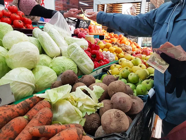 Ο αγοραστής πληρώνει σε μετρητά για αγορασμένα φρούτα κοντά στον πάγκο λαχανικών. Πουλάω σοδειές στην αγορά. Βιταμίνες και μέταλλα για χορτοφάγους. Υγιεινά τρόφιμα και ενεργός τρόπος ζωής. Εμπόριο στην Ουκρανία εθνικού νομίσματος Royalty Free Εικόνες Αρχείου