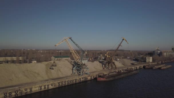 桟橋の近くの海岸に大規模なポートクレーン大規模なヒープに川の砂のバージをアンロードします バルク貨物の輸送のための列車 貨物港の仕事 空中写真ドローン — ストック動画