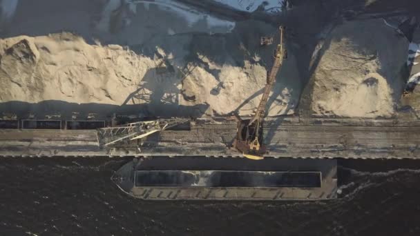 Duże żurawie portowe na brzegu w pobliżu molo rozładowują barkę z piaskiem rzecznym na dużą stertę. Pociąg do przewozu ładunków masowych. Praca w porcie towarowym. Czworośmigłowiec do fotografii lotniczej — Wideo stockowe