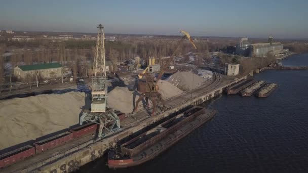 Stora hamnkranar på stranden nära piren lastar av flodens sandpråm på en stor hög. Tåget för transport av bulklast. Lasthamnens arbete. Flygfoto drönare fyrhjuling — Stockvideo