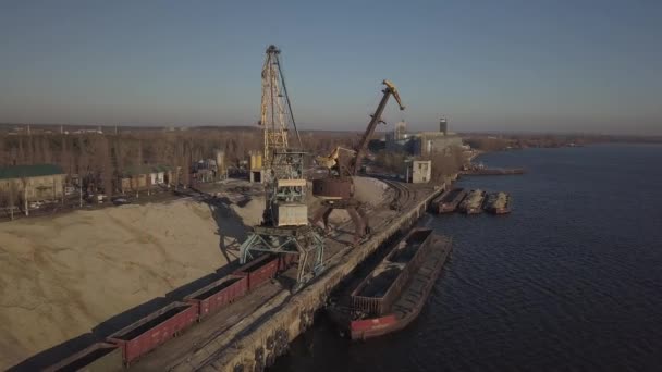 Duże żurawie portowe na brzegu w pobliżu molo rozładowują barkę z piaskiem rzecznym na dużą stertę. Pociąg do przewozu ładunków masowych. Praca w porcie towarowym. Czworośmigłowiec do fotografii lotniczej — Wideo stockowe